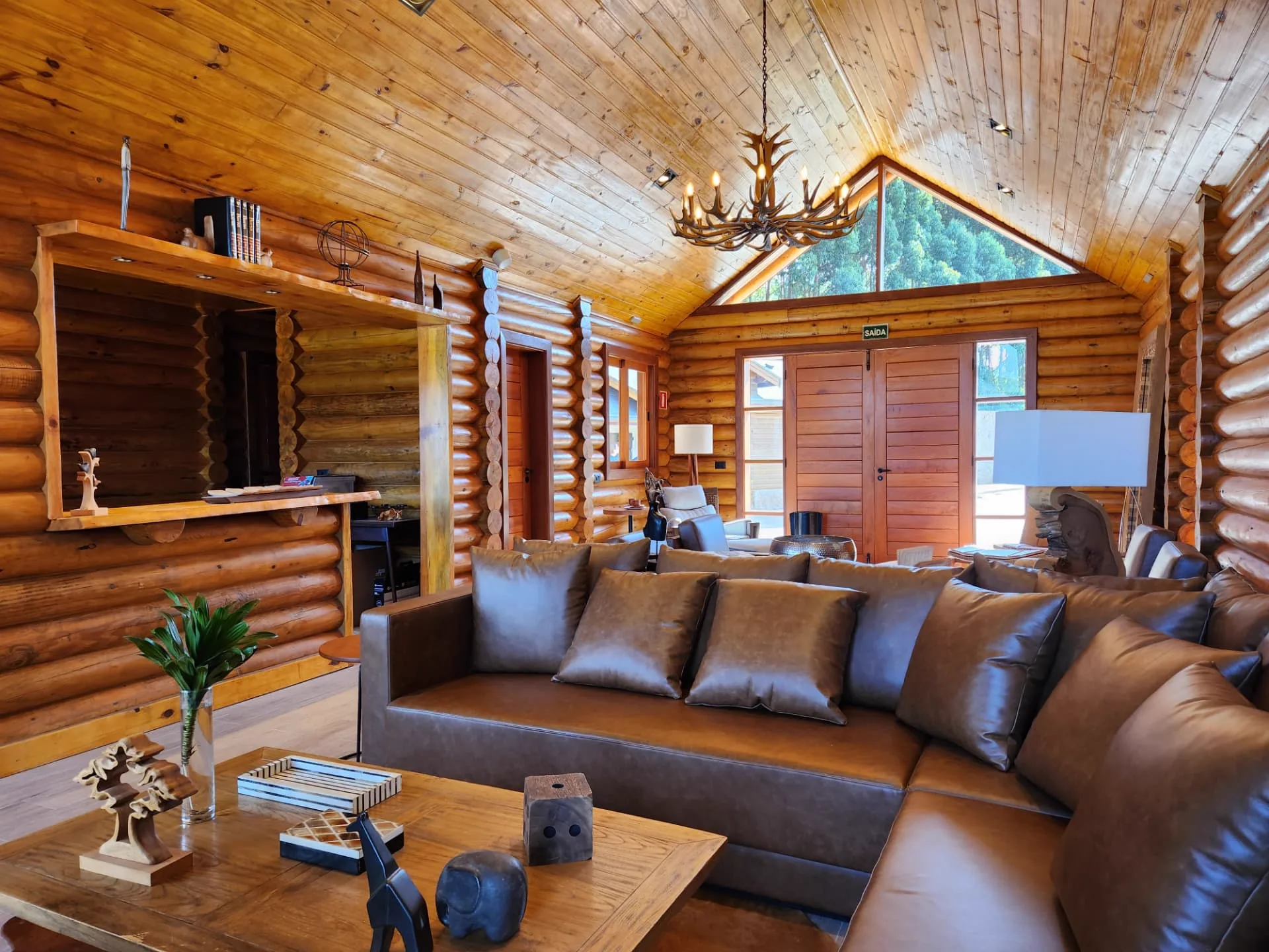 Salão da Log Home da Pousada Maple com troncos aparentes, lareira e vista para a mata